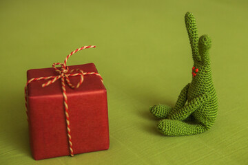 Green crochet bunny sitting far from red wrapped gift box tied with rope bow on light green...