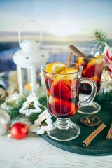 Mulled wine with orange and spices, Christmas decor and burning candles, on a white wooden table against the background of winter nature.
