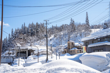 A peaceful winter village nestled in a snow-covered mountain landscape. Thick layers of snow blanket rooftops, streets, and trees under a bright blue sky, capturing the quiet charm of a serene 