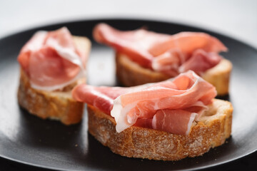 ciabatta with thin prosciutto on black plate on conctere background
