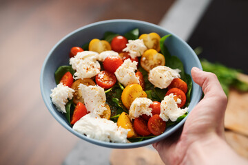 amn hand hold salad with red and yellow cherry tomatoes , mozzarella and spinach in blue bowl