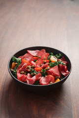 Salad with kale, bresaola and cherry tomatoes decorated with capers in black bowl on walnut wood table