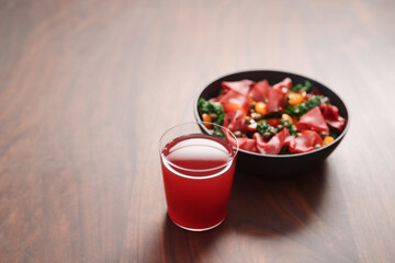 Salad with kale, bresaola and cherry tomatoes decorated with capers in black bowl and glass of red drink on walnut wood table