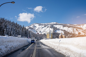 A lone traveler walks along a snowy mountain road under a vivid blue sky. Towering peaks and...