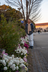 Obraz premium A young woman stands beside a colorful flower garden in a city park during golden hour, surrounded by blooming florals, tall trees, and soft evening light with a historic building in the background.