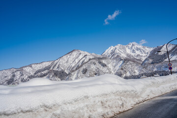 A lone traveler walks along a snowy mountain road under a vivid blue sky. Towering peaks and...