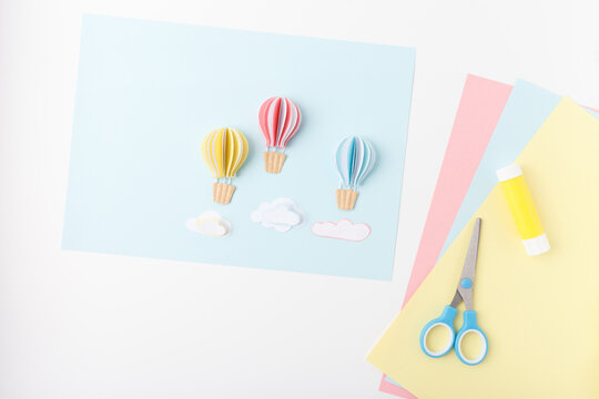 Paper craft scene with colorful balloons, clouds, scissors, glue, pastel sheets on blue background. Ideal for kids crafts, art supply stores, education materials, hobby blogs