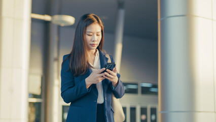 Young Asian businesswoman in formal outfit using smartphone outdoors during evening. Professional appearance in modern urban environment.