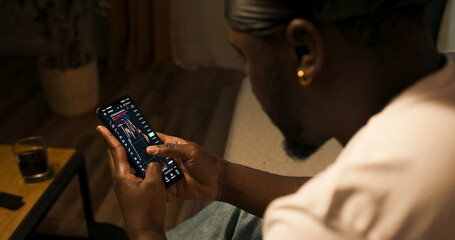 Businessman checks cryptocurrency charts on a mobile app while viewing trading graphs on a laptop. Over-shoulder view of financial market analysis in real time.