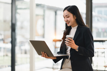 Young Asian businesswoman, smiling and holding coffee cup, works on laptop in modern office with bright natural light, expressing confidence and productivity