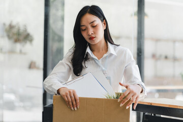 Young Asian woman in white shirt packing office supplies into cardboard box, looking sad and thoughtful, possibly after job loss or resignation, in modern office setting