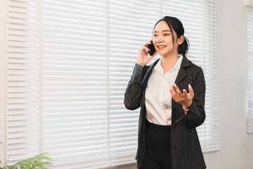 Businesswoman office communication phone conversation professional smiling standing window blinds emotionally expressing confidence while talking on mobile phone in modern office setting