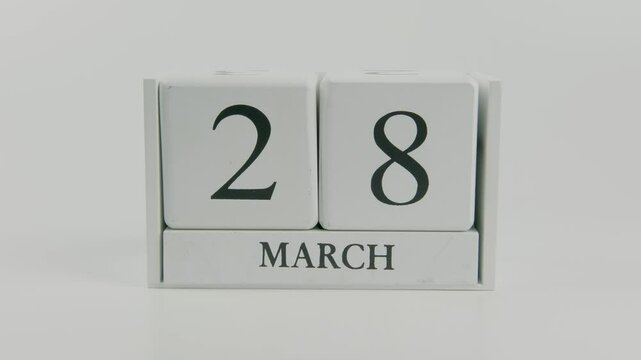 A hand flips a wooden calendar from March 28th to March 29th on a white background, signaling the change in days and the ongoing passage of time