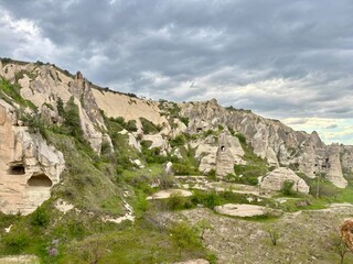 Unique rock formations rise from lush green valleys in Goreme, Cappadocia, showcasing the region's natural beauty under a cloudy sky and hinting at history and adventure