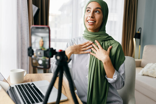 Indoor image with selective focus on lifestyle blogger in hijab discussing cultural aspects on camera with her followers, recording video blog,gesticulating expressing her feelings sitting at table
