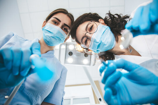 Dentalcare procedure at dentist office. Bottom view of team women dentists in blue surgical mask holding tools and looking at patient's teeth. Patient point of view to dentists