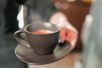 A close-up of a hand holding a black ceramic cup with steaming hot coffee. The blurred background enhances the warm and inviting atmosphere, highlighting the rich aroma of the beverage.