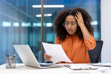 A businesswoman expresses worry while looking at a document. She is seated at a desk with a laptop, papers, and a pen holder in front of her.