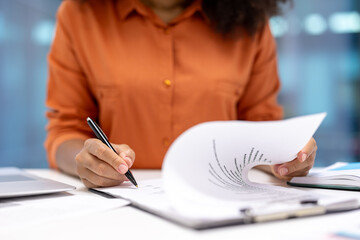 A person in an orange shirt is writing on a document in an office setting, focusing on work with...