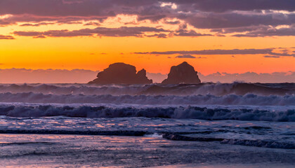 Obraz premium Seashore Sunset: The fiery embrace of the setting sun casts a radiant glow upon the rhythmic ocean waves and dramatic rock formations, offering a captivating coastal panorama.
