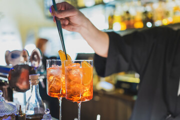 A bartender garnishes two Aperol Spritz cocktails with orange slices using tongs. The drinks, served in tall glasses filled with ice cubes, create a refreshing and vibrant scene at the bar.