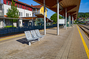 An empty train station platform featuring sleek metal benches and wooden canopies. The platform is well-maintained, with a yellow safety line and tactile paving for accessibility