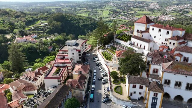 Aerial 4K footage of the National Palace of Sintra, Portugal. Smooth drone flight over historic white building with iconic chimneys, green hills, and traditional architecture