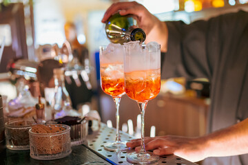 Bartender pouring sparkling wine into two glasses with ice and orange cocktail, creating refreshing drinks at a lively bar counter.