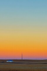 Fototapeta premium Sunrise over a vast, desolate plain. A small, simple building sits on the horizon. Pale, blurred colors fill the sky