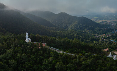 A serene aerial view of a large white Buddha statue perched on a forested hill, surrounded by lush greenery and overlooking a peaceful valley with distant villages. The tranquil setting exudes spiritu