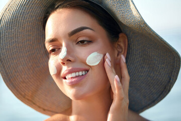 Smiling woman applies sunscreen on her face while wearing a large sun hat, enjoying a sunny beach...
