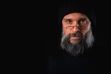emotional, facial, angry portrait of a bearded man in a black hat and glasses looking into the camera. Dramatic portrait in subdued lighting, expressive face, emotional tension, dark background