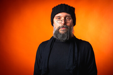 Portrait of a stylish mature man with a beard and glasses wearing a black hat and clothes, standing against a vibrant orange background. Trendy, confident, modern look.