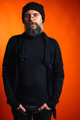 Portrait of a stylish mature man with a beard and glasses wearing a black hat and clothes, standing against a vibrant orange background. Trendy, confident, modern look.