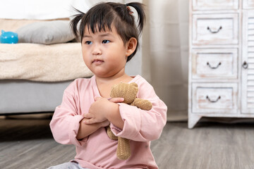 Asian toddler girl wearing pink shirt sitting on wooden floor holding brown stuffed teddy bear...