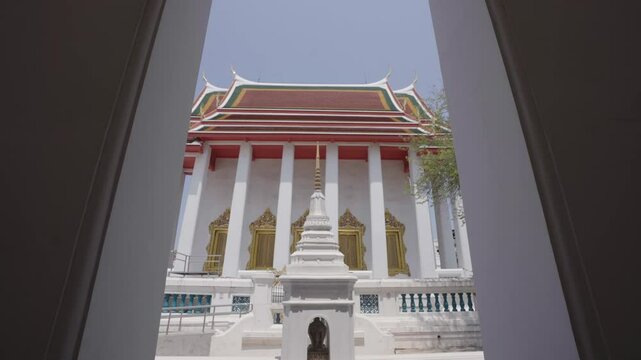A low angle view of a traditional Thai Buddhist temple hall with white pillars and golden doors, standing beside a historic white prang pagoda against a clear sky.