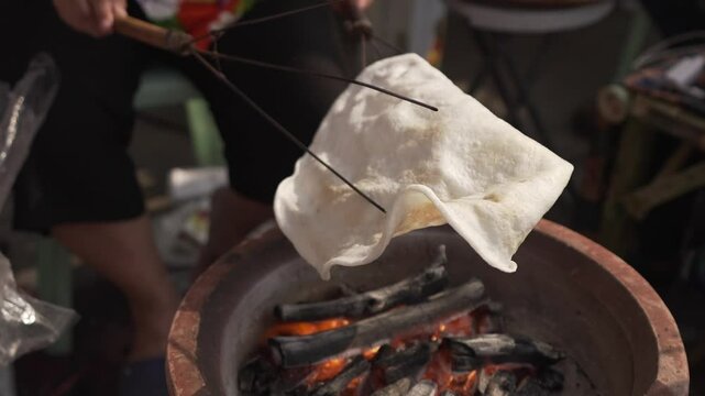 An elderly vendor skillfully flips a giant rice cracker over hot charcoal using a wooden tong.