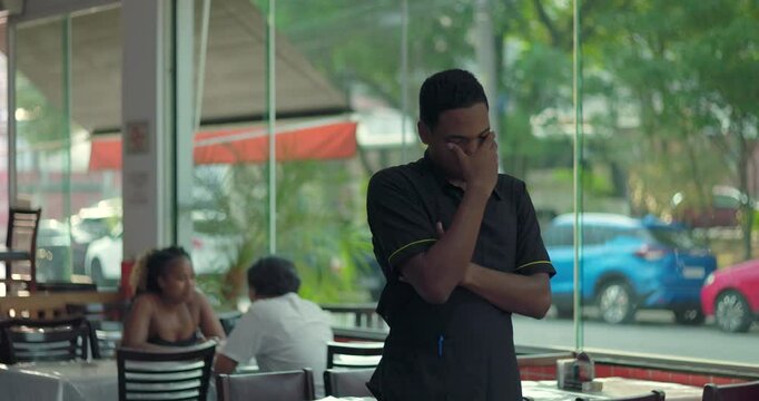 Restaurant worker standing with hand over eyes, showing fatigue or stress during shift, indoor dining area visible in background near window