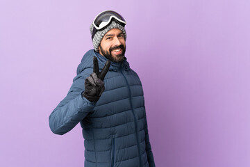 Skier man with snowboarding glasses over isolated purple background smiling and showing victory sign