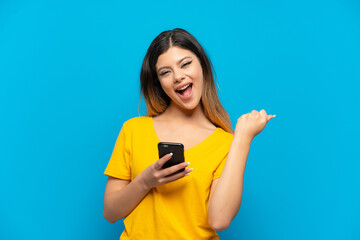 Fototapeta premium Young Russian girl isolated on blue background with phone in victory position