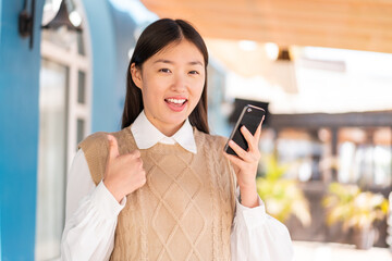 Young Chinese woman at outdoors using mobile phone while doing thumbs up