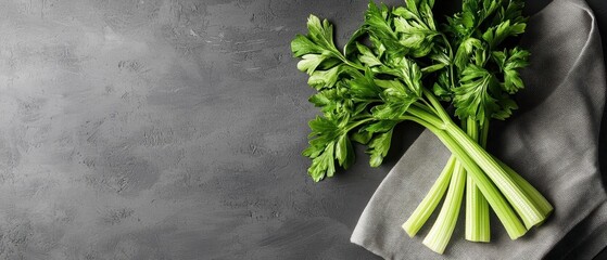 Bunch of fresh green celery leaves on a dark grey textured background. the celery is lying on a grey cloth napkin on the right side of the image.