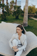 Woman in light suit sitting outside on abstract stone bench with contemplative expression in spring...