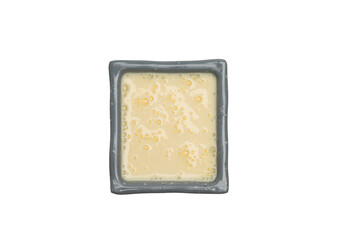 A square grey dish holds a creamy, pale yellow liquid with small bubbles, possibly a sauce, dip, or melted substance.
