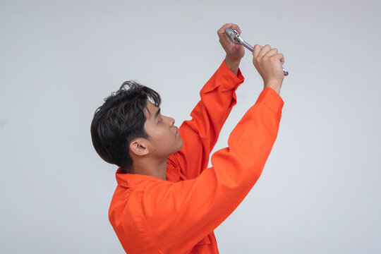 Side view of a young Southeast Asian male mechanic in orange coveralls using a wrench, simulating repair on overhead machinery. Isolated on a light background.