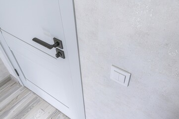 close-up shot of a white door with a dark handle next to a light switch on a textured, light gray wall. The floor is visible at the bottom left, featuring light wood planks.