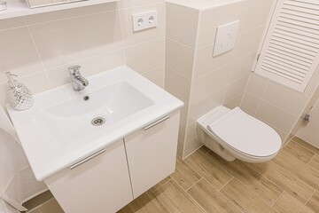 Clean, modern bathroom interior with white fixtures, light beige tiles, and wood-like flooring....