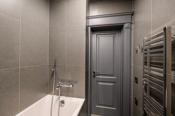 Modern bathroom interior featuring gray tile walls, a white bathtub, a gray door with trim, and a chrome towel warmer.