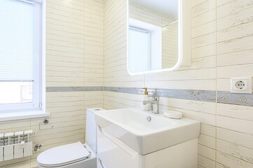 bright, modern bathroom features off-white wood-look tiles, a white sink and toilet, and a window with blinds. A blue decorative tile border adds a subtle accent.