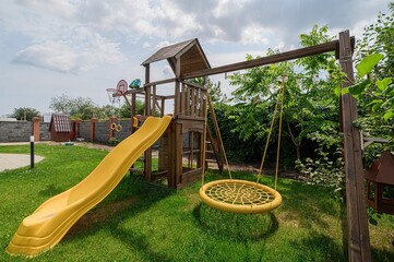 vibrant playground with a wooden play structure, yellow slide, and round swing set on a green lawn under a partly cloudy sky.